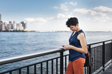 Seitenansicht einer Frau, die ihr Smartphone benutzt, während sie auf einer Promenade am Fluss gegen den Himmel in der Stadt steht - CAVF62645