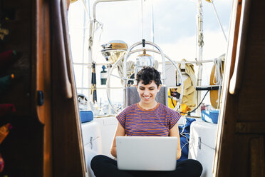 Smiling woman using laptop computer while sitting in boat seen through window - CAVF62652