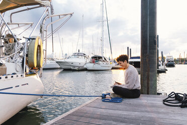 Side view of woman writing while sitting on pier at harbor against cloudy sky during sunset - CAVF62662