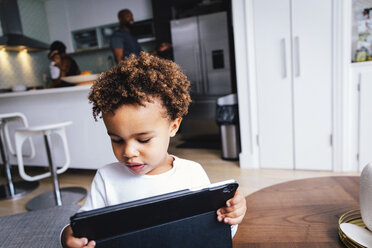 Nahaufnahme eines süßen Jungen, der einen Tablet-Computer benutzt, während er zu Hause vor seiner Familie steht - CAVF62669