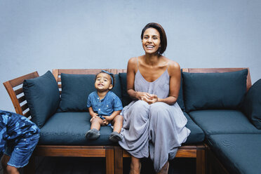 Portrait of happy mother with cute daughter sitting on sofa against wall at home - CAVF62678