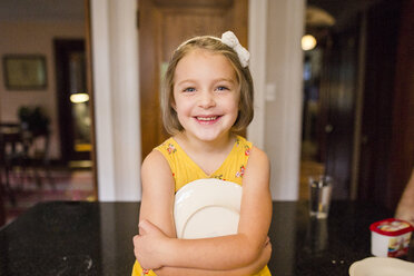 Porträt eines süßen lächelnden Mädchens, das einen Teller hält, während es zu Hause am Esstisch sitzt - CAVF62681