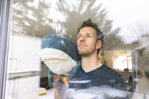 Man looking out of window, carrying toy shark on his shoulder - JOSF03121
