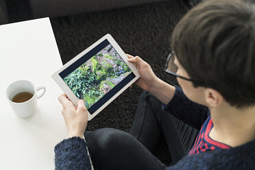 Frau sitzt zu Hause auf dem Boden und betrachtet ein Foto auf einem digitalen Tablet - SBOF01817