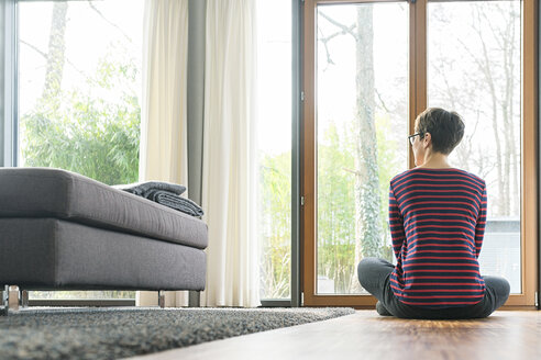 Back view of woman sitting on the floor of living room looking out of window - SBOF01827