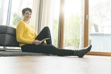 Portrait of happy woman with digital tablet sitting on the floor of living room - SBOF01833