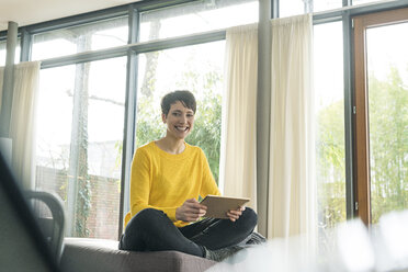 Porträt einer glücklichen Frau mit digitalem Tablet, die auf der Couch im Wohnzimmer sitzt - SBOF01834