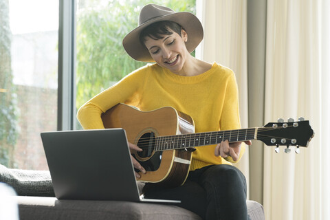 Portrait of smiling woman sitting on couch with laptop playing guitar stock photo
