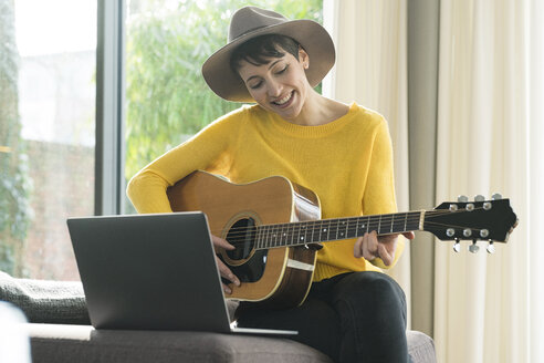 Porträt einer lächelnden Frau, die mit einem Laptop auf der Couch sitzt und Gitarre spielt - SBOF01844