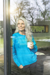 Porträt einer zufriedenen reifen Frau mit einer Tasse Kaffee, die an der Terrassentür lehnt und in die Ferne schaut - SBOF01851