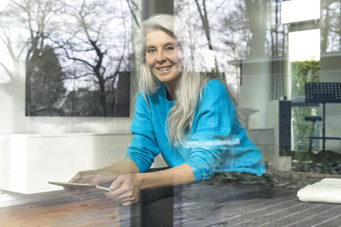 Porträt einer lächelnden reifen Frau mit digitalem Tablet, die zu Hause auf der Couch sitzt und aus dem Fenster schaut - SBOF01855