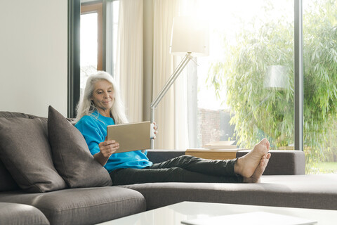 Porträt einer zufriedenen reifen Frau, die zu Hause auf der Couch sitzt und auf ein digitales Tablet schaut, lizenzfreies Stockfoto