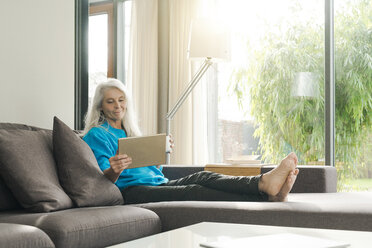 Porträt einer zufriedenen reifen Frau, die zu Hause auf der Couch sitzt und auf ein digitales Tablet schaut - SBOF01856
