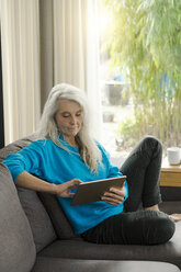 Portrait of mature woman sitting on the couch at home using digital tablet - SBOF01858