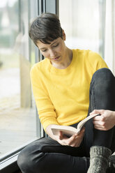 Frau sitzt auf dem Boden neben der Terrassentür und liest ein Buch - SBOF01866