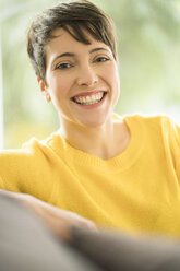 Portrait of happy woman with short brown hair wearing yellow pullover sitting on the couch at home - SBOF01873