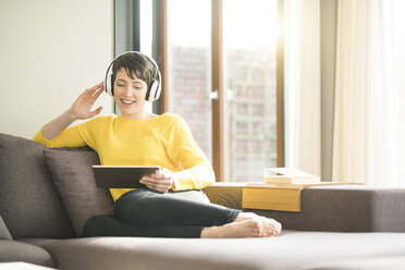 Portrait of happy woman sitting on the couch at home listening music with headphones while looking at tablet - SBOF01875