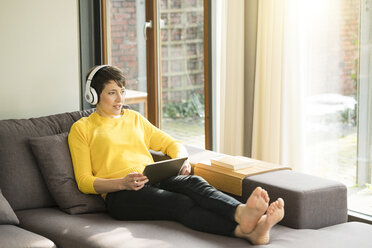 Porträt einer Frau mit digitalem Tablet, die zu Hause auf der Couch sitzt und mit Kopfhörern Musik hört - SBOF01876