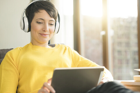 Porträt einer glücklichen Frau mit Kopfhörern, die zu Hause auf der Couch sitzt und auf ein digitales Tablet schaut, lizenzfreies Stockfoto