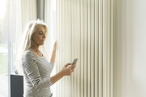 Mature woman with smartphone checking heating in living room of smart home - SBOF01886