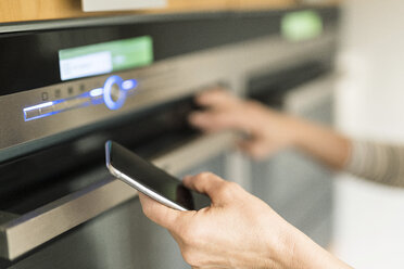 Hand of woman with smartphone checking oven in kitchen at her smart home - SBOF01888