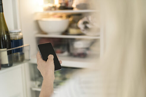 Hand of woman with smartphone checking fridge in kitchen at smart home - SBOF01890