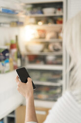 Hand einer Frau mit Smartphone, die den Kühlschrank in der Küche eines Smart Home überprüft - SBOF01891
