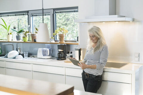 Zufriedene Frau steht in der Küche und benutzt ein digitales Tablet, lizenzfreies Stockfoto