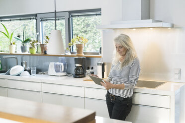 Zufriedene Frau steht in der Küche und benutzt ein digitales Tablet - SBOF01893