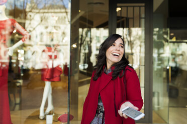 Happy woman holding cell phone in front of shop window - VABF02233