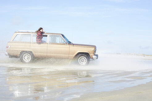 Germany, St Peter-Ording, girl leaning out of window of off-road vehicle driving through water on the beach - AMEF00033