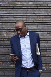 Portrait of smiling businessman with newspaper wearing glasses and blue suit looking at mobile phone - IGGF00909