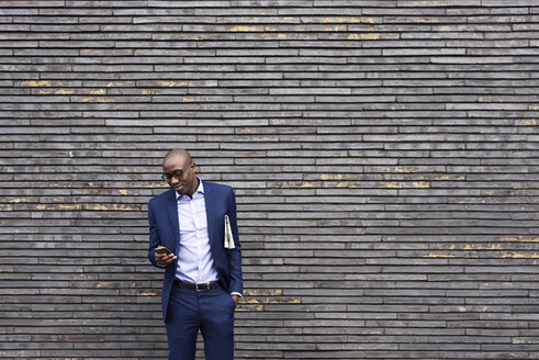 Portrait of smiling businessman with newspaper wearing glasses and blue suit looking at mobile phone - IGGF00911