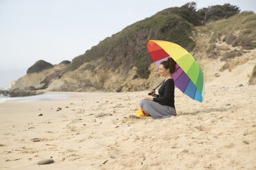 Frau mit buntem Regenschirm sitzt am Strand - KBF00560