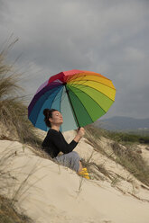 Frau mit buntem Sonnenschirm am Strand sitzend, Sonnenbad nehmend - KBF00570