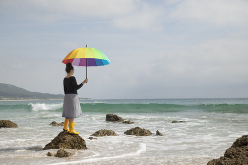 Woman with colorful umbrella standing on a stone at the beach - KBF00571