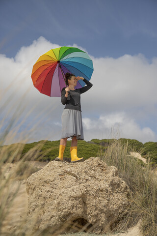 Frau mit buntem Regenschirm steht auf einem Felsen und schaut in die Ferne, lizenzfreies Stockfoto