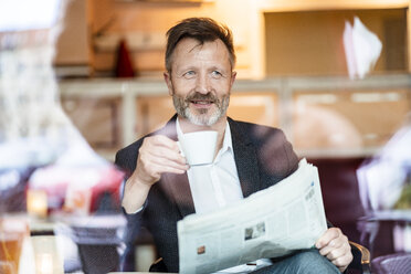 Portrait of smiling mature businessman with newspaper sitting in a coffee shop drinking coffee - DIGF06005