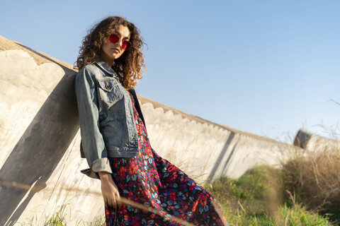 Porträt einer jungen Frau mit lockigem braunem Haar und roter Sonnenbrille, die an einer Wand lehnt, lizenzfreies Stockfoto