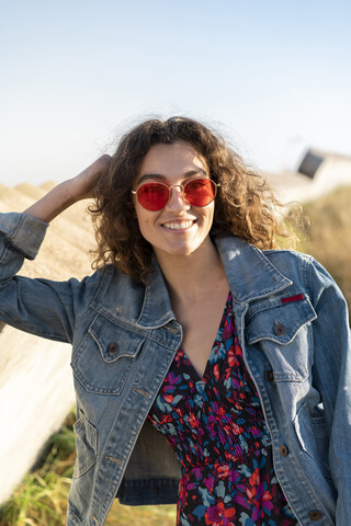 Porträt einer glücklichen jungen Frau mit lockigem braunem Haar und roter Sonnenbrille, die sich an eine Wand lehnt, lizenzfreies Stockfoto