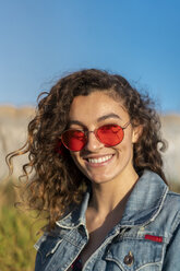 Portrait of happy young woman with curly brown hair wearing red sunglasses - AFVF02575
