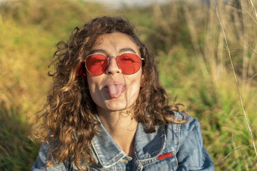 Porträt einer jungen Frau mit lockigem braunem Haar und roter Sonnenbrille, die die Zunge herausstreckt - AFVF02577