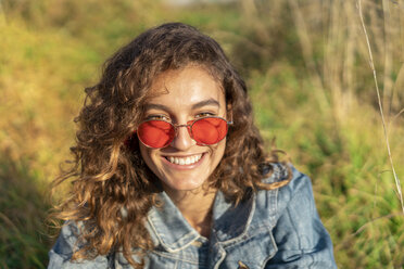 Porträt einer glücklichen jungen Frau mit lockigem braunem Haar und roter Sonnenbrille - AFVF02578