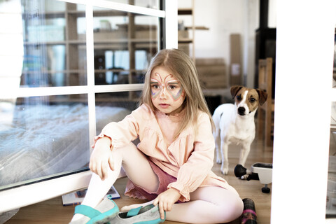 Blondes Mädchen als Schmetterling geschminkt, auf dem Boden sitzend, Jack Russel Terrier im Hintergrund, lizenzfreies Stockfoto