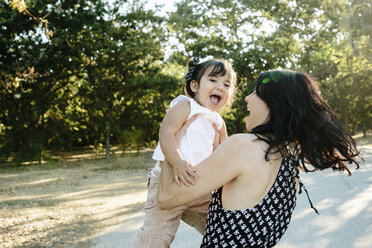 Side view of happy mother carrying cute daughter while standing on road against trees in park - CAVF62715