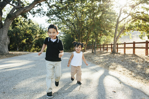 Portrait of happy cute siblings running on road against trees in park stock photo