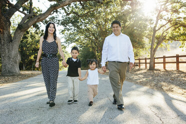 Porträt einer glücklichen Familie, die sich an den Händen hält, während sie auf der Straße gegen Bäume im Park läuft - CAVF62722