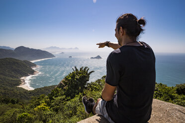 Tourist, der auf das Meer zeigt, während er auf einer Klippe gegen den blauen Himmel an einem sonnigen Tag sitzt - CAVF62781