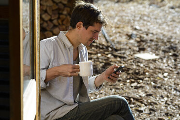 Mann, der eine Kaffeetasse hält und sein Smartphone benutzt, während er an einem sonnigen Tag im Wald sitzt - CAVF62786