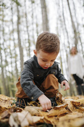 Cute son crouching on dry leaves while mother standing in background at forest during autumn - CAVF62804